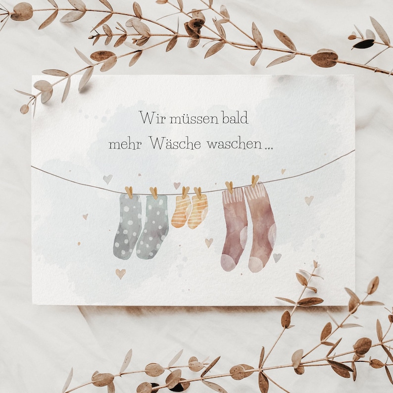 May include: A rectangular greeting card featuring a watercolour illustration of a clothesline with two grey polka-dot socks, two small orange striped socks, and two brown socks. The text reads: Wir m&uuml;ssen bald mehr W&auml;sche waschen...