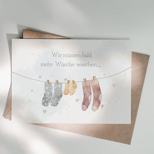May include: A white greeting card featuring a watercolour illustration of a clothesline with two grey polka-dot socks, two small yellow striped socks, and two brown socks. The text reads: Wir m&uuml;ssen bald mehr W&auml;sche waschen...