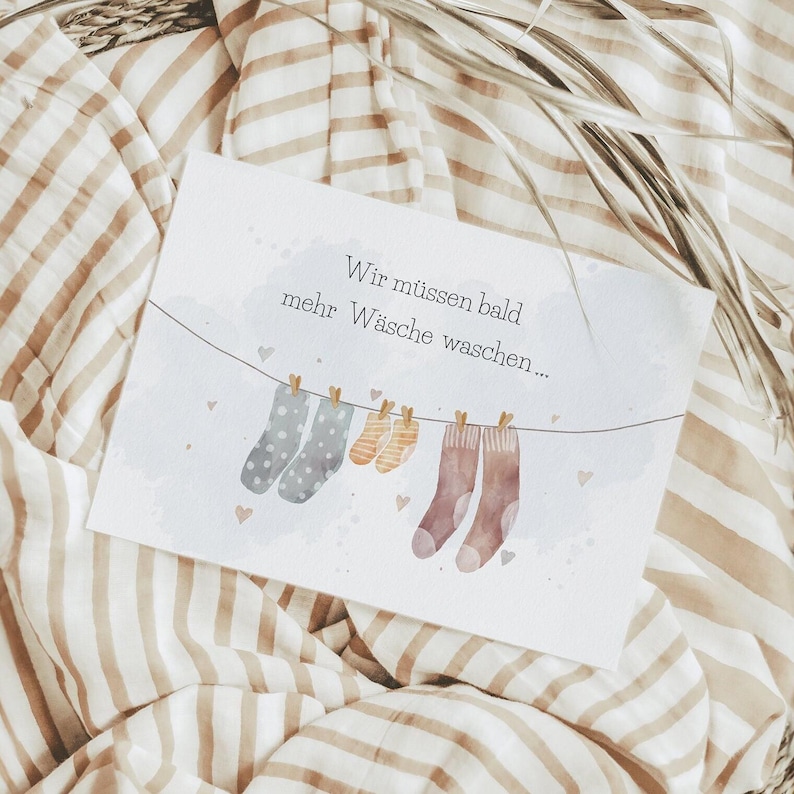 May include: A rectangular white card featuring a watercolour illustration of a clothesline with two grey polka-dot socks, two small yellow striped socks, and two brown socks hanging from wooden clips, with the German text Wir m&uuml;ssen bald mehr W&auml;sche waschen.