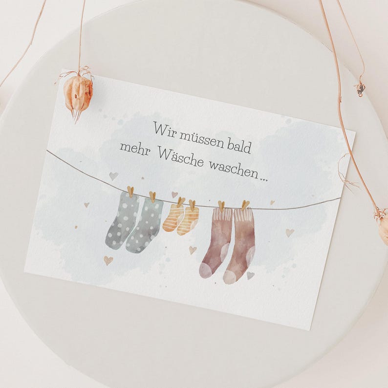 May include: A rectangular card featuring a watercolour illustration of three pairs of socks hanging on a clothesline with wooden clips. The text reads Wir m&uuml;ssen bald mehr W&auml;sche waschen. The background is white with light blue watercolour accents.