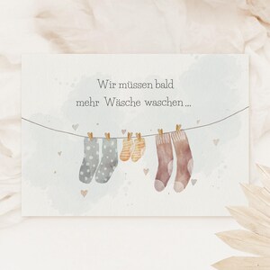May include: A rectangular card featuring a watercolour illustration of a clothesline with three pairs of socks in varying sizes and patterns, including polka dots and stripes, with the German text Wir m&uuml;ssen bald mehr W&auml;sche waschen.