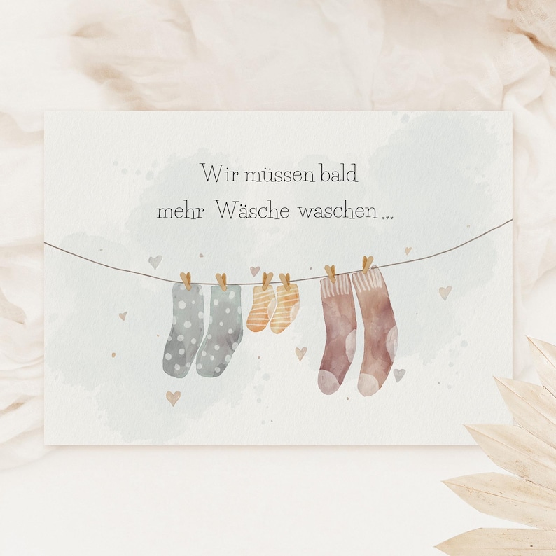 May include: A rectangular card featuring a watercolour illustration of a clothesline with three pairs of socks in varying sizes and patterns, including polka dots and stripes, with the German text Wir m&uuml;ssen bald mehr W&auml;sche waschen.