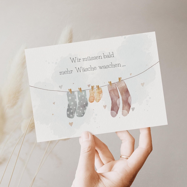 May include: A rectangular greeting card featuring a watercolour illustration of a clothesline with two grey polka-dot socks, two small orange striped baby socks, and two brown socks. The text reads: Wir m&uuml;ssen bald mehr W&auml;sche waschen...