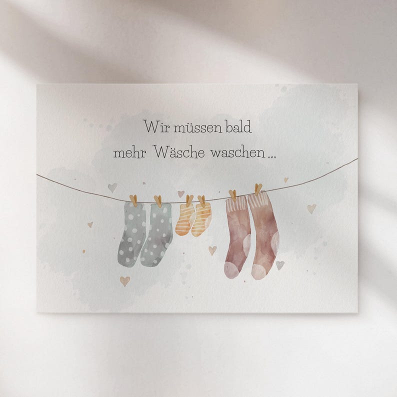 May include: A rectangular greeting card featuring a watercolour illustration of a clothesline with two grey polka-dot socks, two small yellow striped baby socks, and two brown socks hanging from wooden clips, with the text Wir m&uuml;ssen bald mehr W&auml;sche waschen.