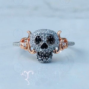 May include: A silver ring with a skull-shaped design set with black and white diamonds. The skull has a detailed face with black eyes and a diamond-encrusted smile. The ring band is adorned with delicate, swirling details.