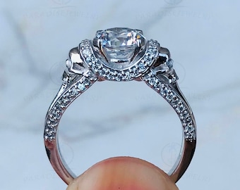 Puede incluir: Un anillo de compromiso de plata con una gran piedra central redonda y transparente. El anillo tiene un diseño de calavera a cada lado de la piedra central y está engastado con piedras transparentes más pequeñas.
