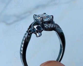 May include: A black skull ring with a large, clear, square-shaped gemstone. The ring is made of metal and has a twisted band.