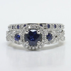 May include: A set of two rings. The engagement ring features a large, round, dark blue gemstone surrounded by smaller white gemstones. The wedding band is a thin band with small blue gemstones. Both rings are silver in color.
