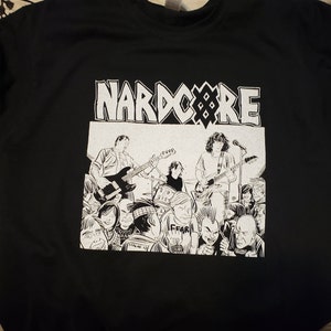May include: Black t-shirt with a white graphic print of a band called "Nardcore". The graphic features a band playing on stage with a crowd of people in front of them.