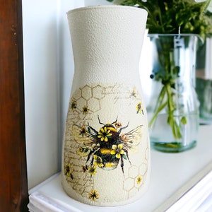 May include: A white ceramic vase with a textured surface features a bee and honeycomb design. The vase is decorated with yellow flowers and a vintage-style script.
