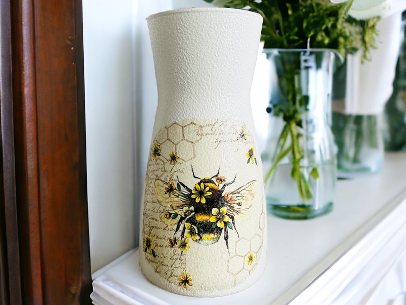 K&ouml;nnte beinhalten: Eine wei&szlig;e Keramikvase mit strukturierter Oberfl&auml;che zeigt ein Bienen- und Wabenmuster. Die Vase ist mit gelben Blumen und einer Schrift im Vintage-Stil verziert.