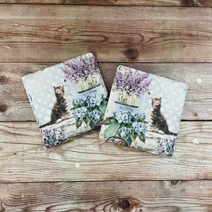May include: Two square decorative pillows with a cat and floral design. Each pillow features a tabby cat, blue hydrangeas, and lavender in a blue pot with the word "FLOWERS". The pillows are on a wooden surface.