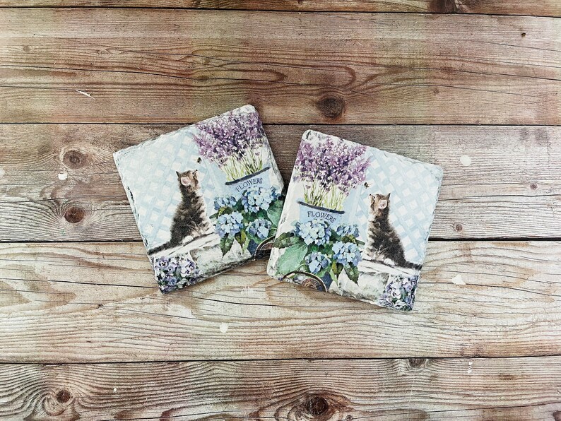 May include: Two square decorative pillows with a cat and floral design. Each pillow features a tabby cat, blue hydrangeas, and lavender in a blue pot with the word "FLOWERS". The pillows are on a wooden surface.