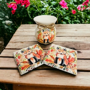 May include: Two ceramic coasters with a fox design. The coasters are painted with a fox, leaves, and berries in shades of orange, red, and green. A white ceramic vase with a fox design is also pictured.