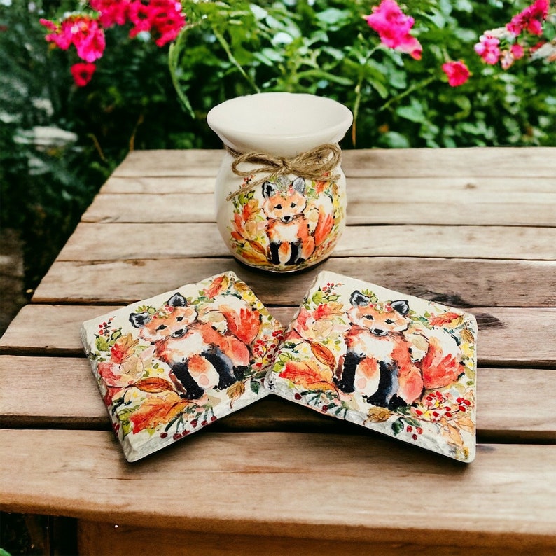May include: Two ceramic coasters with a fox design. The coasters are painted with a fox, leaves, and berries in shades of orange, red, and green. A white ceramic vase with a fox design is also pictured.