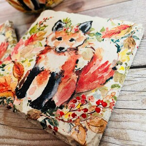 May include: A square coaster with a watercolor design of a fox in a forest setting. The fox is orange and white with black markings. The background is a light beige with red berries, green leaves, and yellow flowers.