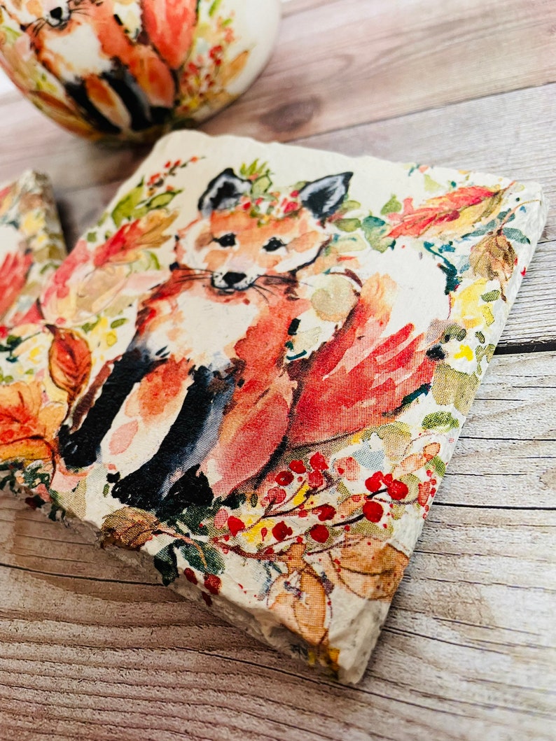 May include: A square coaster with a watercolor design of a fox in a forest setting. The fox is orange and white with black markings. The background is a light beige with red berries, green leaves, and yellow flowers.