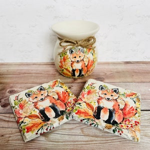May include: Two ceramic coasters with a fox design. The coasters feature a fox with orange fur, a white chest, and black legs. The fox is surrounded by autumn leaves and berries. The coasters are set on a wooden surface.