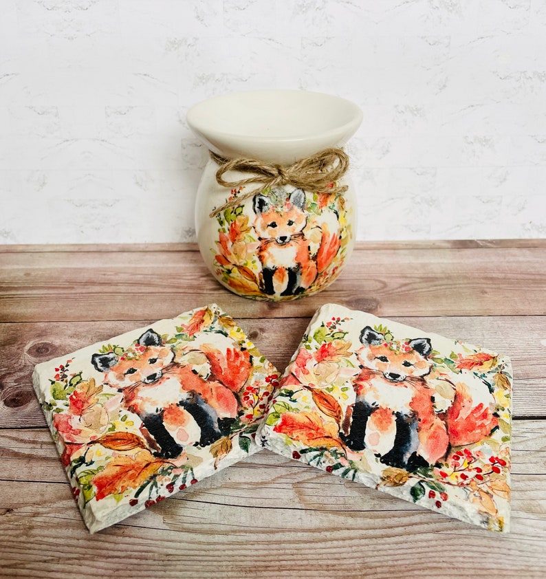 May include: Two ceramic coasters with a fox design. The coasters feature a fox with orange fur, a white chest, and black legs. The fox is surrounded by autumn leaves and berries. The coasters are set on a wooden surface.