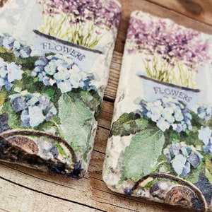 May include: Two decorative stone coasters with a floral design. Each coaster features a blue flower pot with the word "FLOWERS" and a cluster of blue hydrangeas. Lavender flowers are in the background. A cat is visible on the right coaster.