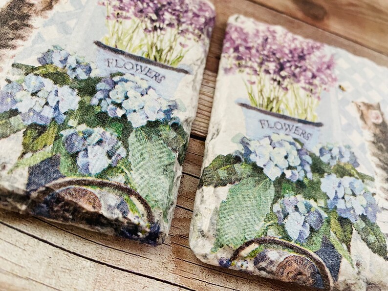 May include: Two decorative stone coasters with a floral design. Each coaster features a blue flower pot with the word "FLOWERS" and a cluster of blue hydrangeas. Lavender flowers are in the background. A cat is visible on the right coaster.