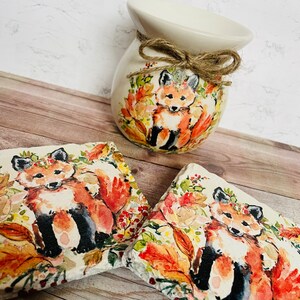 May include: A white ceramic wax warmer with a brown twine bow and a fox design. The warmer is surrounded by two square coasters with a fox design. The coasters are made of slate and have a watercolor-like design.