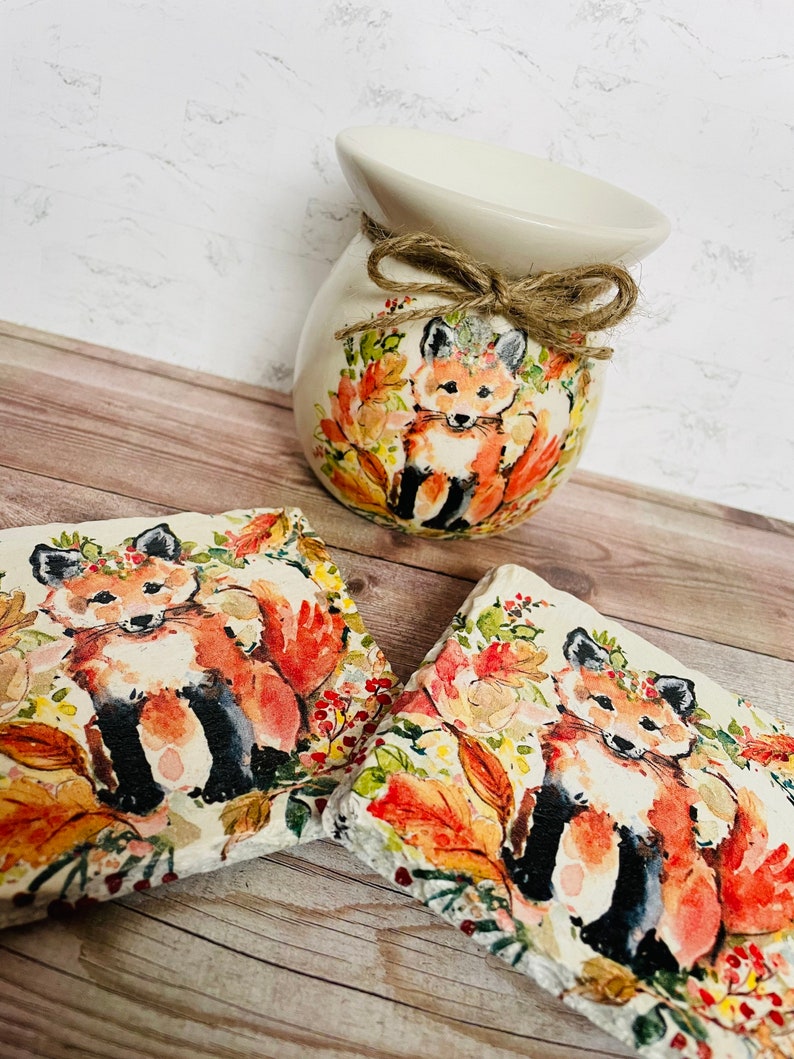May include: A white ceramic wax warmer with a brown twine bow and a fox design. The warmer is surrounded by two square coasters with a fox design. The coasters are made of slate and have a watercolor-like design.