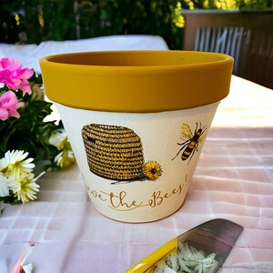 May include: A yellow and white ceramic flower pot with a bee and honeycomb design. The pot has the text "Save the Bees!" written on it.
