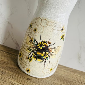 K&ouml;nnte beinhalten: Wei&szlig;e Keramikvase mit einem Bienen- und Wabenmuster. Die Biene ist von gelben Blumen umgeben. Die Vase hat eine strukturierte Oberfl&auml;che.