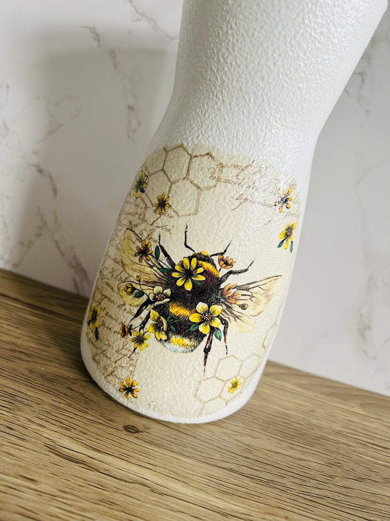 K&ouml;nnte beinhalten: Wei&szlig;e Keramikvase mit einem Bienen- und Wabenmuster. Die Biene ist von gelben Blumen umgeben. Die Vase hat eine strukturierte Oberfl&auml;che.