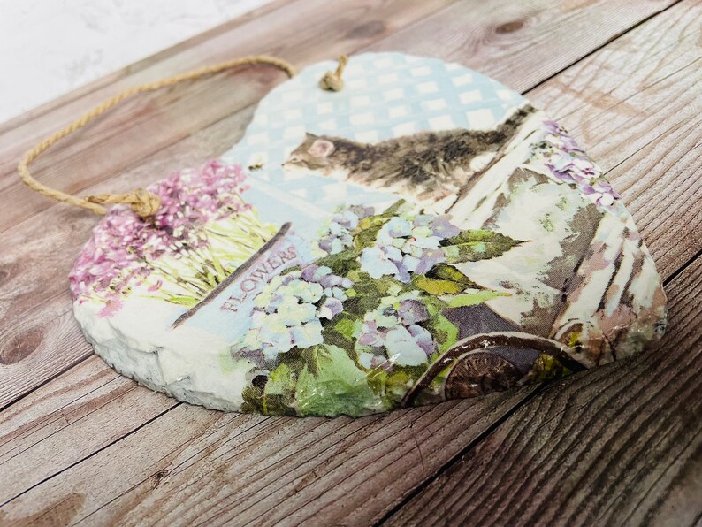 K&ouml;nnte beinhalten: Eine herzf&ouml;rmige Schiefertafel mit einem Blumendesign und dem aufgedruckten Wort "FLOWERS". Die Tafel hat einen rustikalen, Vintage-Look und ist mit einem blau-wei&szlig; karierten Muster, einer Katze und einer Schubkarre voller Blumen verziert.