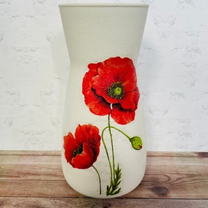 May include: White ceramic vase with a floral design featuring red poppies. The vase has a textured surface and a wide mouth.