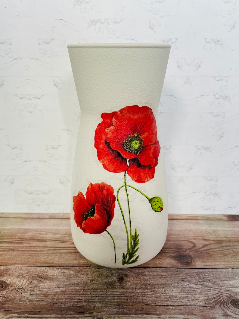 May include: White ceramic vase with a floral design featuring red poppies. The vase has a textured surface and a wide mouth.