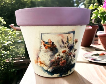May include: A white ceramic flower pot with a lavender rim. The pot features a watercolor image of a cat looking out a window with flowers in the background.