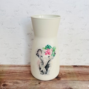 May include: White ceramic vase with a floral design featuring an elephant wearing a flower crown. The vase has a textured surface.