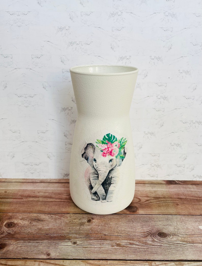 May include: White ceramic vase with a floral design featuring an elephant wearing a flower crown. The vase has a textured surface.