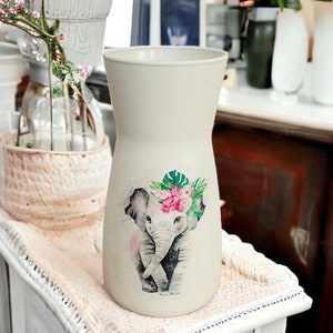 May include: A white ceramic vase with a textured surface features a floral design with a pink and green elephant. The elephant is wearing a floral crown.