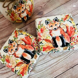 May include: Two square stone coasters with a watercolor design of foxes in a forest setting. The foxes are orange and white with black accents. The background features fall leaves in shades of orange, yellow, and red.