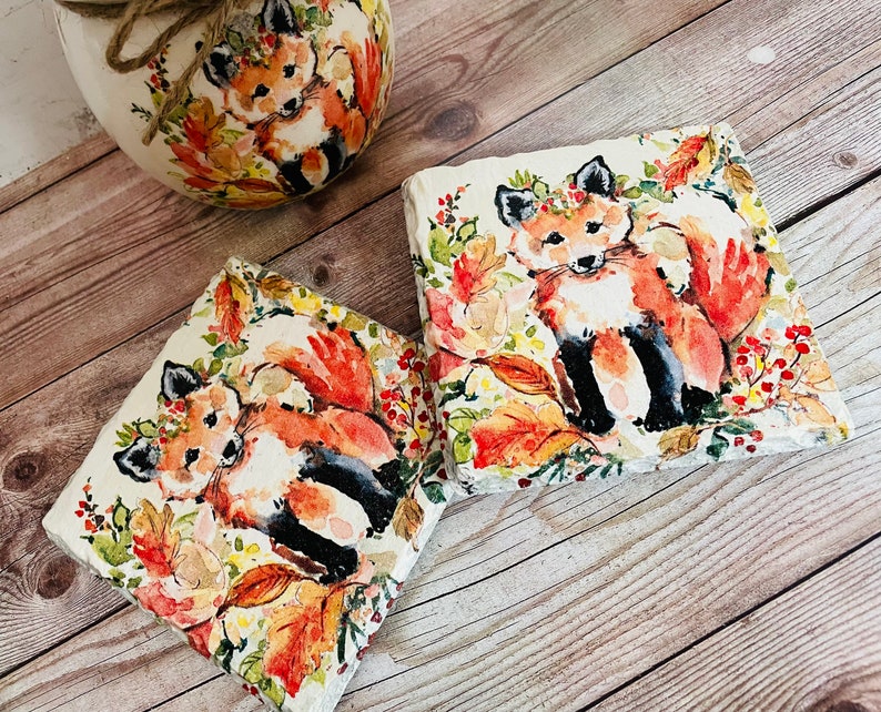 May include: Two square stone coasters with a watercolor design of foxes in a forest setting. The foxes are orange and white with black accents. The background features fall leaves in shades of orange, yellow, and red.