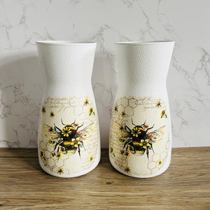 K&ouml;nnte beinhalten: Zwei wei&szlig;e Keramikvasen mit einem Bienen- und Wabenmuster. Die Vasen sind mit einer gelben und schwarzen Biene, gelben Blumen und einem Wabenmuster verziert.