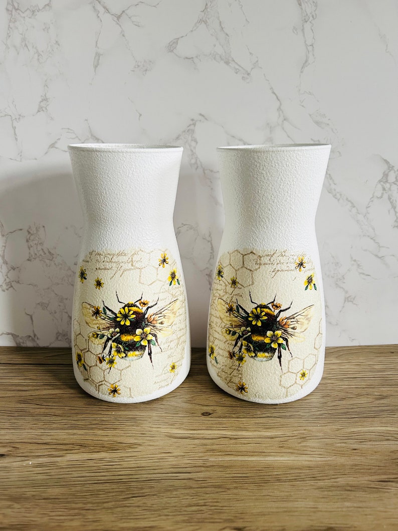 K&ouml;nnte beinhalten: Zwei wei&szlig;e Keramikvasen mit einem Bienen- und Wabenmuster. Die Vasen sind mit einer gelben und schwarzen Biene, gelben Blumen und einem Wabenmuster verziert.