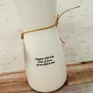 May include: White ceramic vase with a brown twine bow. The vase has the text "Happy 18th Isla, Tons of love. From Gill & Ian" printed on the side.