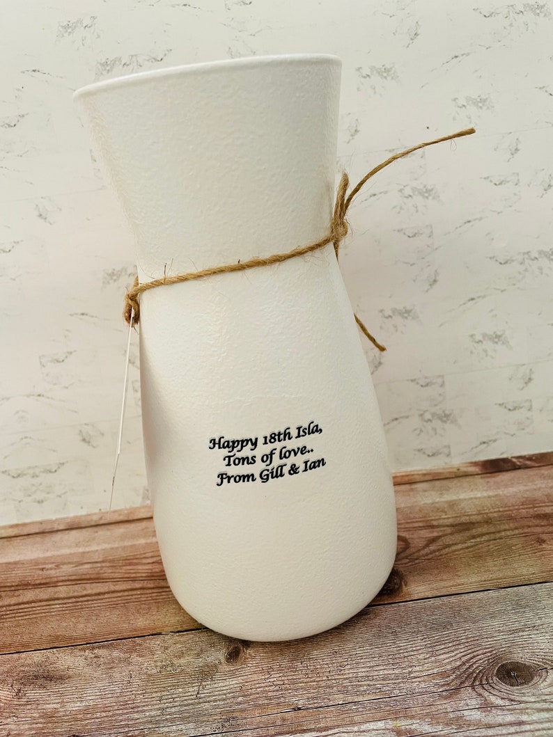 May include: White ceramic vase with a brown twine bow. The vase has the text "Happy 18th Isla, Tons of love. From Gill & Ian" printed on the side.