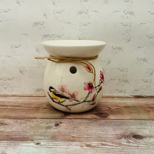 May include: White ceramic wax melt burner with a floral design featuring a small bird and pink flowers. The burner has a brown twine tied around the top.