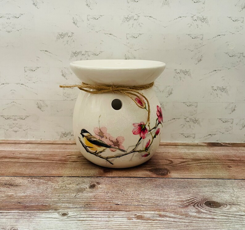 May include: White ceramic wax melt burner with a floral design featuring a small bird and pink flowers. The burner has a brown twine tied around the top.