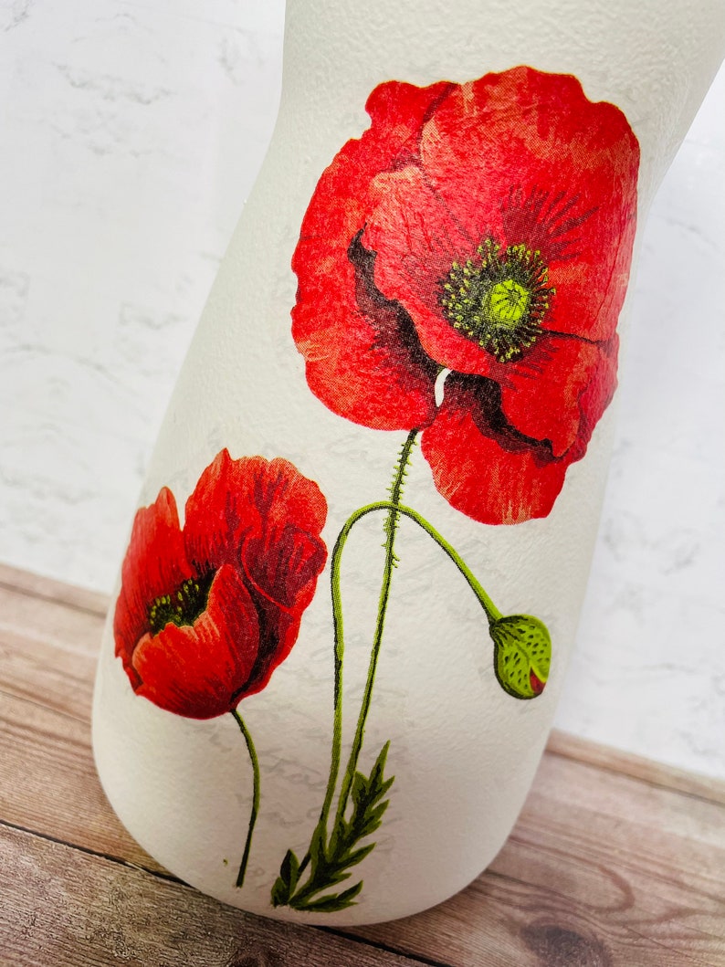 May include: White ceramic vase with a red poppy flower design. The vase has a textured surface and a narrow neck.