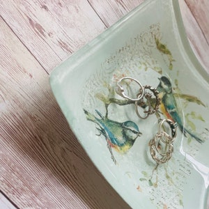 May include: A light blue glass dish with a white background and a floral design. The dish has two bluebirds and several silver rings inside.