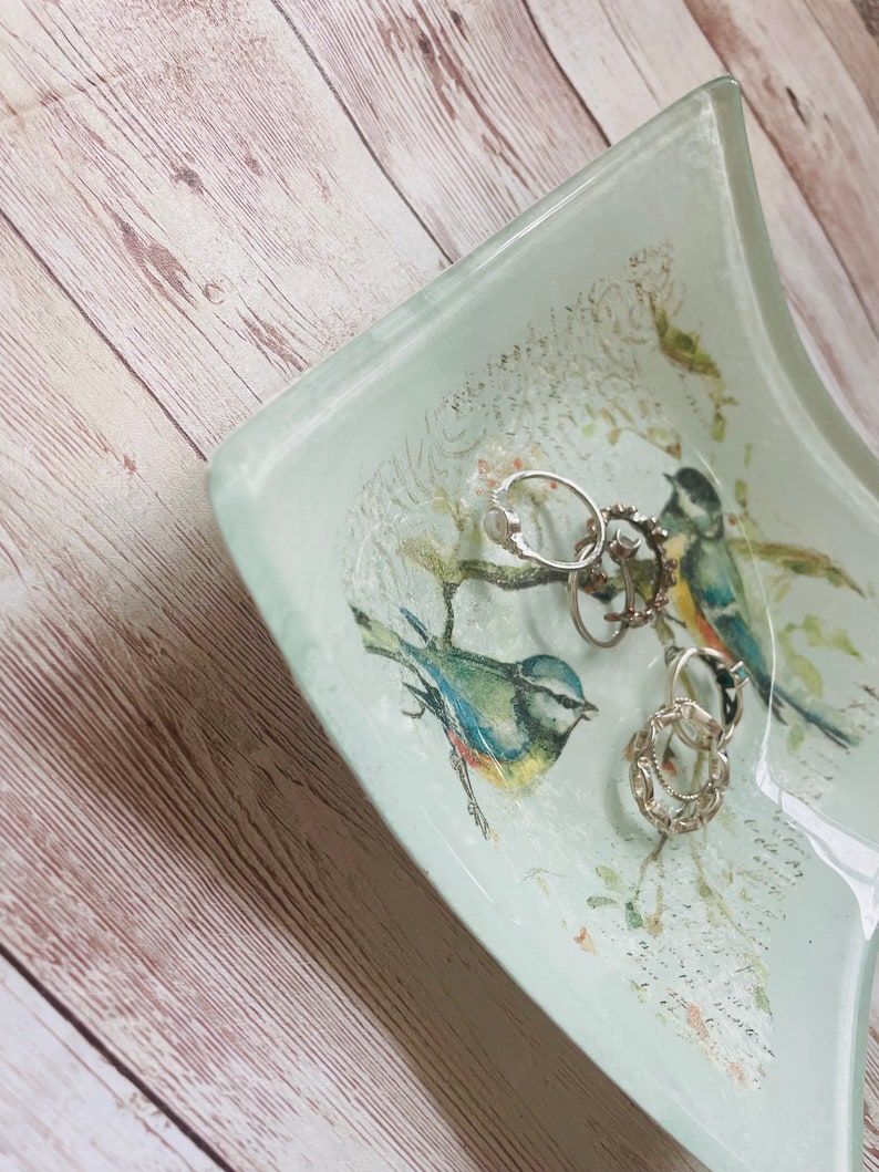 May include: A light blue glass dish with a white background and a floral design. The dish has two bluebirds and several silver rings inside.