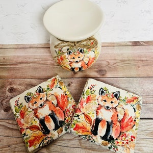 May include: Two ceramic coasters with a watercolor design of a fox in a forest setting. The fox is wearing a flower crown and surrounded by leaves and berries. The coasters are a light brown color and have a slightly textured surface.