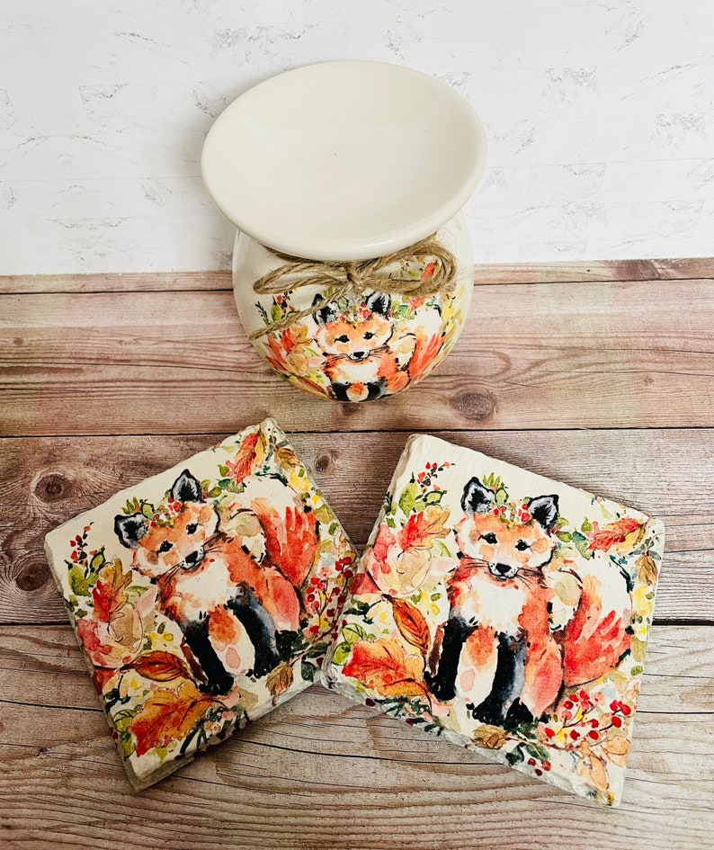 May include: Two ceramic coasters with a watercolor design of a fox in a forest setting. The fox is wearing a flower crown and surrounded by leaves and berries. The coasters are a light brown color and have a slightly textured surface.